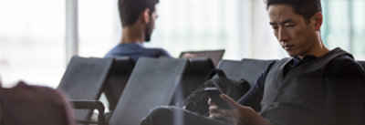 Muškarac sedi u terminalu aerodroma i gleda u telefon. U pozadini sedi druga osoba i radi na laptop računaru.