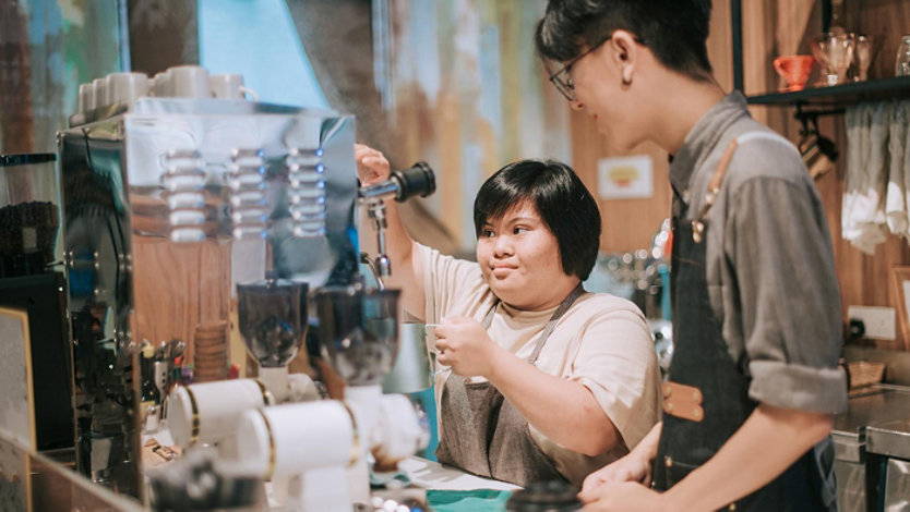 Asian female with a disability learning from barista making coffee in her cafe.