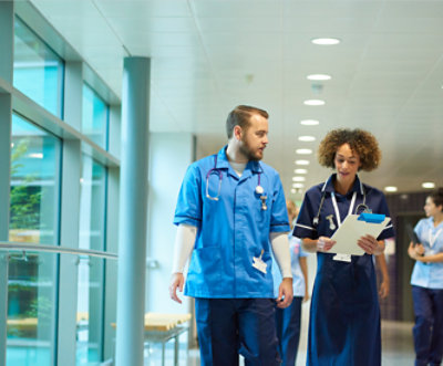 Two medical professionals walking down a hallway having a conversation and looking at a clipboard 