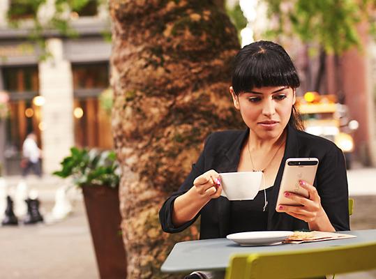 A person sitting at a table outside, sipping a coffee and looking at their mobile phone