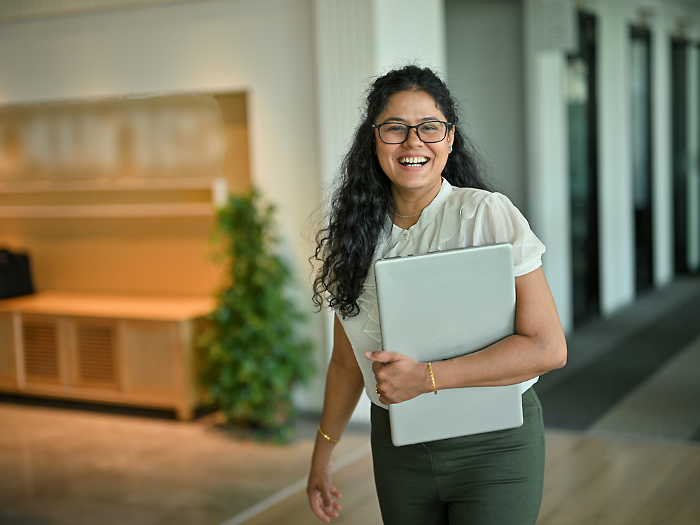 Eine fröhliche Frau mit lockigem Haar, die eine Brille und Geschäftskleidung trägt, hält einen Laptop, während sie in einem Büroflur steht.