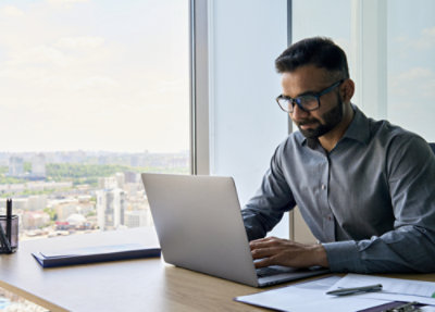 Person sitting at a desk in a modern office, working on a laptop near a large window with a cityscape view in the background.