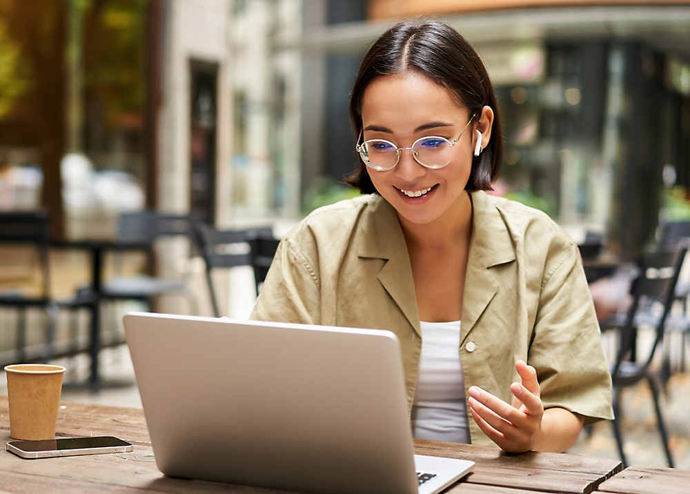 Jonge vrouw met een bril en een smartphone die glimlacht terwijl ze een laptop gebruikt aan een cafétafel buiten