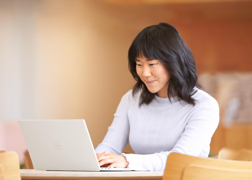A young asian woman smiling while working on a laptop at a wooden table in a well-lit room.