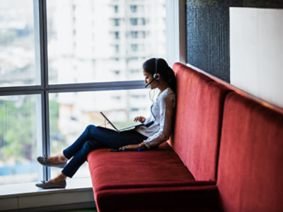 A woman wearing a headset sits on a red couch near a large window, working on a laptop.