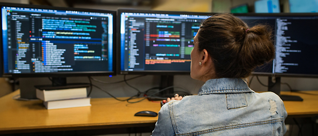 Mujer codificando en varias pantallas de equipos que muestran código de software en una oficina apagada.