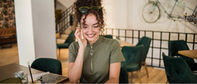 A woman with curly hair in a green shirt is sitting in a cafe, smiling, while talking on a phone.