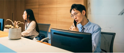 A man in glasses talks on a phone at a computer, while a woman sits beside him working. 