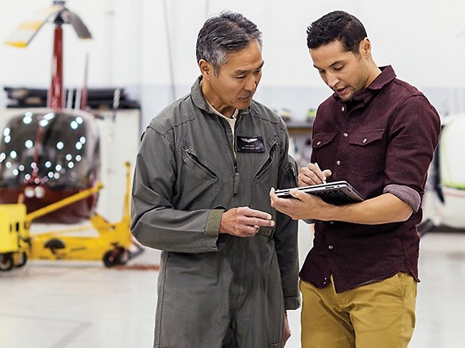 Two men reviewing a document in an aircraft hangar with a helicopter in the background.