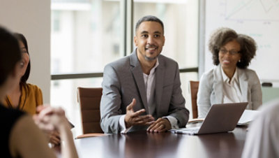 Person in boardroom speaking to others while using a laptop