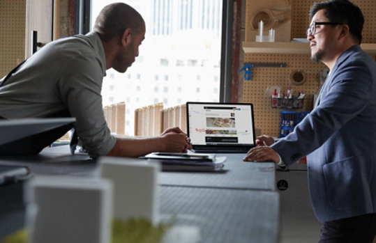 To voksne menn som samarbeider i et arkitektmodellstudio stående på hver sin side av et tegnebord. En mann med en åpen Microsoft Laptop 3 med Microsoft Edge på skjermen