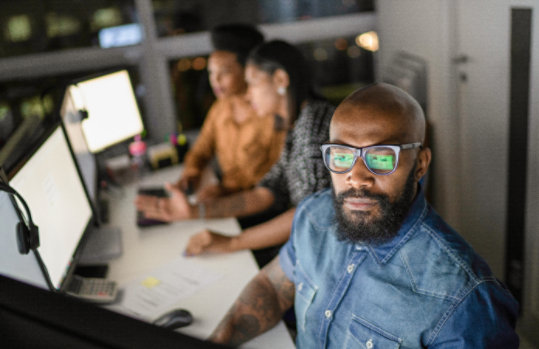 A man sitting in front of monitors with two co-workers in the background