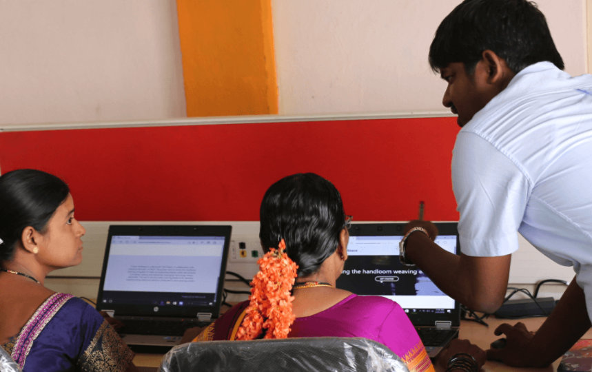 In een computerlokaal spreekt een instructeur met drie studenten die naar hun laptop kijken.
