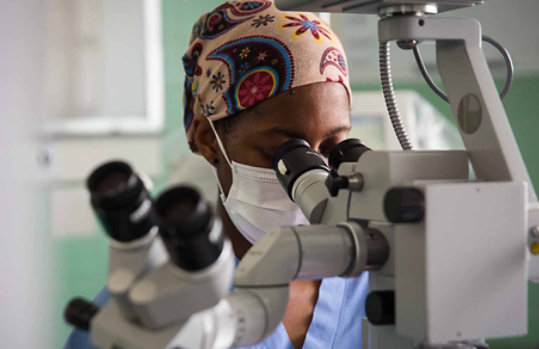 A person wearing a headscarf and medical face mask looks through a microscope