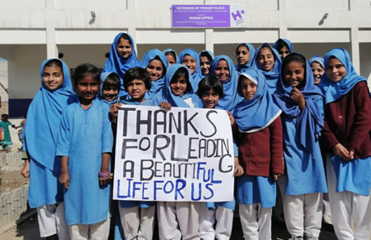 Eine Gruppe von Kindern in Schuluniformen hält ein Schild mit der Aufschrift „Danke, dass du ein schönes Leben für uns führst“