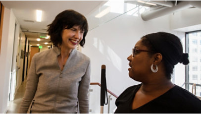 A person using a white cane in a conversation with another person in a hallway.