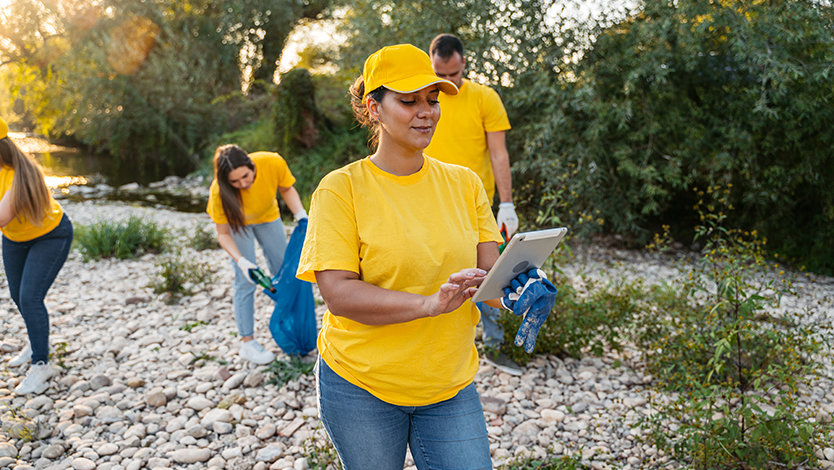 Volunteers in yellow shirts clean up a riverbank while one person uses a tablet.