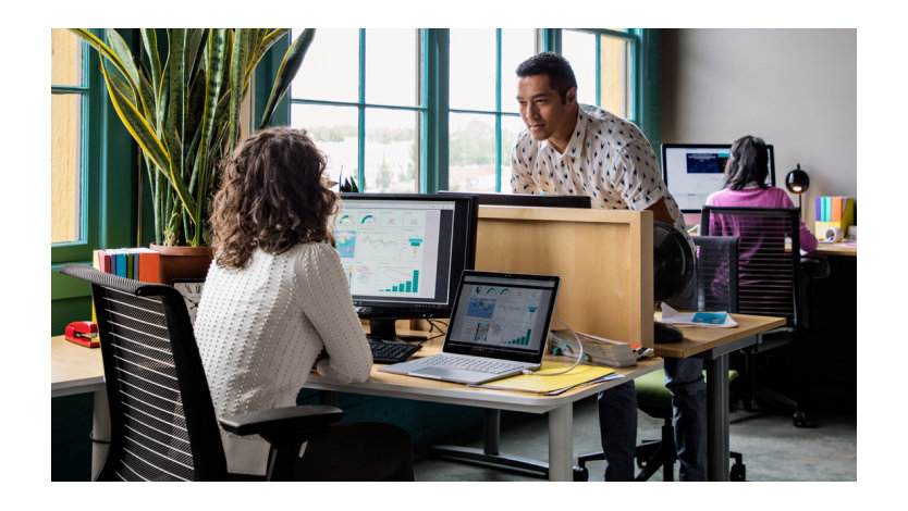 In an office, two coworkers, one in front of the other, talk from cubicle to cubicle.