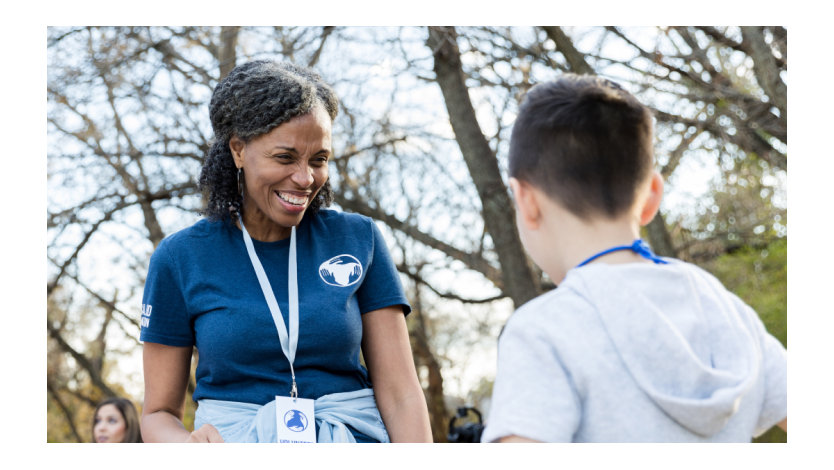 An adult volunteer wearing a lanyard standing outdoors smiling at a child.