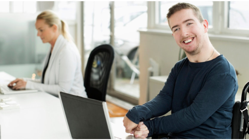 Une personne en fauteuil roulant souriant à la caméra tout en utilisant un ordinateur portable, avec un autre employé dans le bureau en arrière-plan.