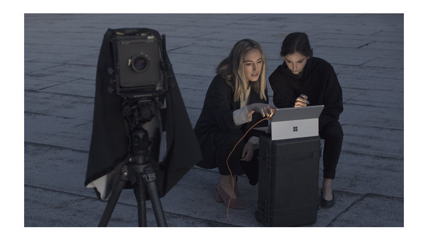 Two people use a laptop, near a large camera on a tripod