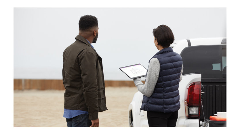 Two adults outside collaborating at a work site. One is holding a black Microsoft Surface Pro X in tablet mode and the other is viewing the screen.