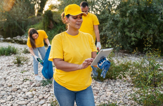 Volunteers in yellow shirts clean up a riverbank while one person uses a tablet.