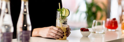 Osoba drži koktel ukrašen limetom i nanom, za belim pultom na kojem se nalaze flaše i mala činija sa malinama