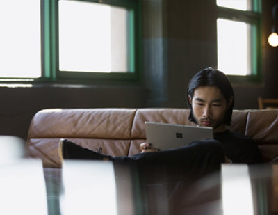 A man with long black hair sitting on a couch using a tablet.