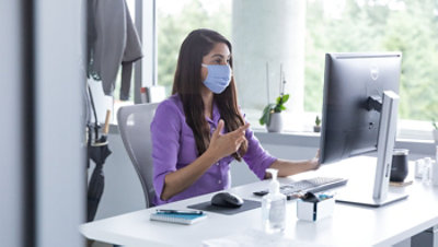 Eine Person mit Maske, die über einen Desktopmonitor im Homeoffice an einem Videochat teilnimmt