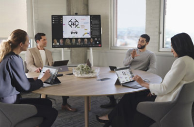 Four people in a meeting room with a Teams Rooms device displaying a Teams meeting and a presentation on a television.
