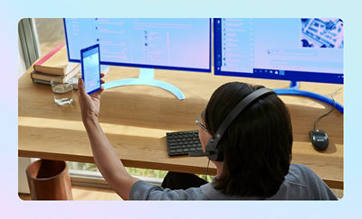 Person using a smartphone and dual monitors at a workstation to manage digital communications and applications