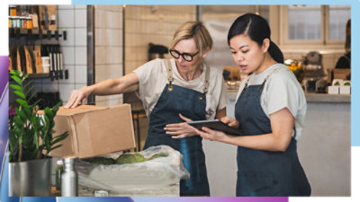 Two workers in aprons reviewing information on a tablet while handling a cardboard box at a work counter.