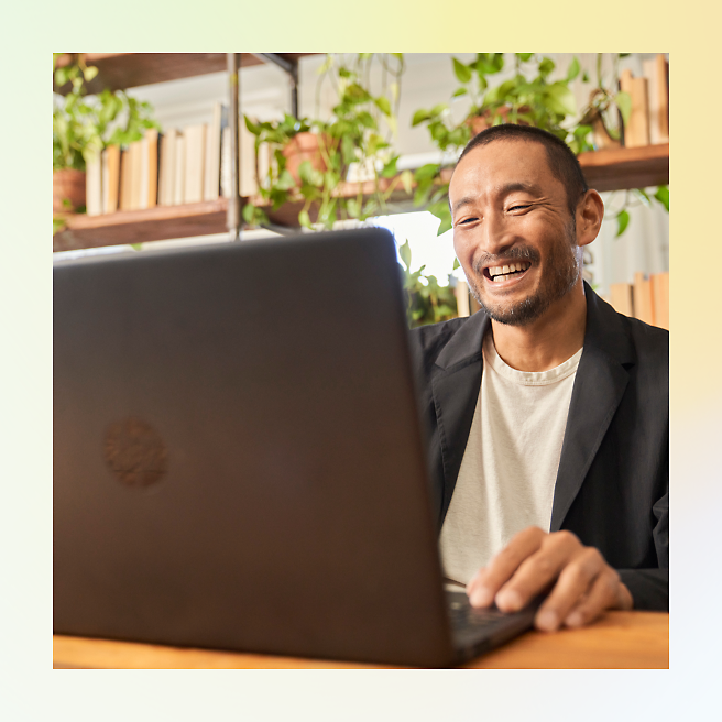 Homem sorrindo enquanto usa um laptop em uma sala com plantas e livros.