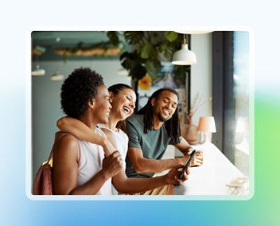 Parlak ışıklı bir kafede gülen ve birlikte akıllı telefona bakan üç arkadaş.