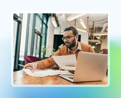 Gözlüklü ve turuncu kazaklı adam, modern bir ofiste dizüstü bilgisayarı olan bir masada belgeleri gözden geçiriyor.