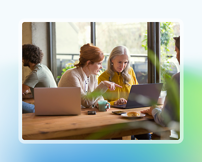 Twee vrouwen die praten naast een laptop in een druk café met een andere persoon die op de achtergrond op een laptop werkt.