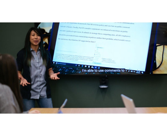 Una mujer realiza una presentación delante de un monitor de gran tamaño que cuelga de una pared. 