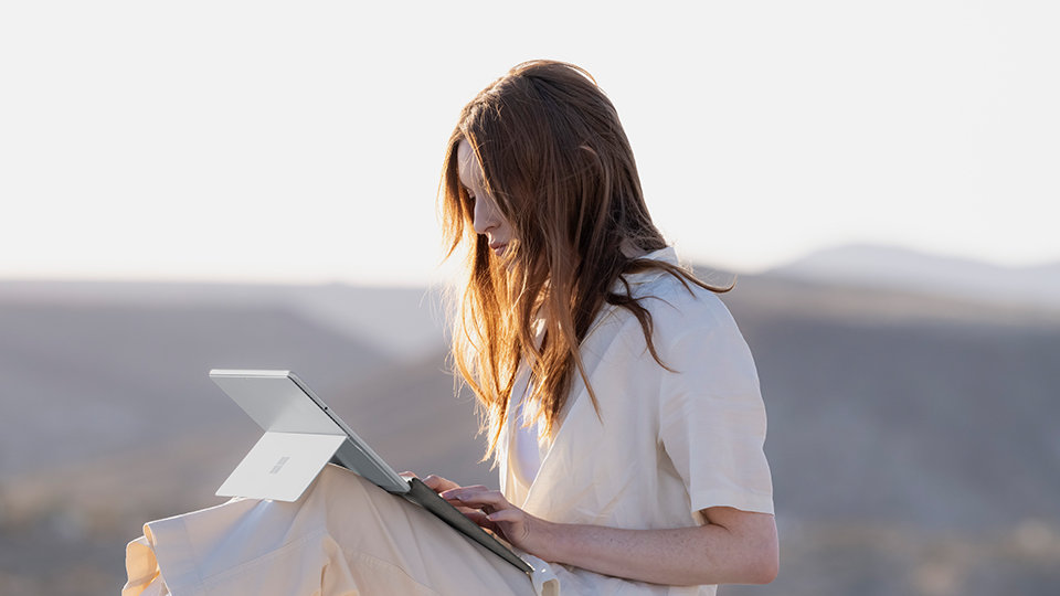A person sitting in the sun with mountains nearby working on a Surface Pro Signature Keyboard for Business.