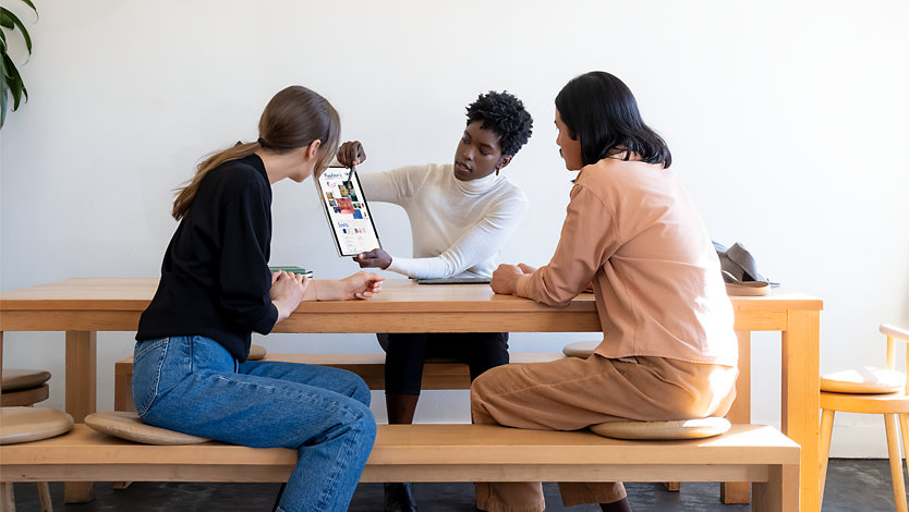 Une personne utilisant un stylet Slim Pen et un appareil Surface pour montrer son travail à deux autres personnes assises à une table en bois.
