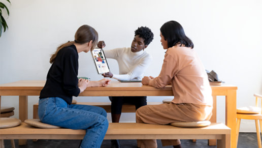 Una persona usando un Slim Pen 2 y un dispositivo Surface para enseñarle su trabajo a otras dos personas que están sentadas en una mesa de madera. 