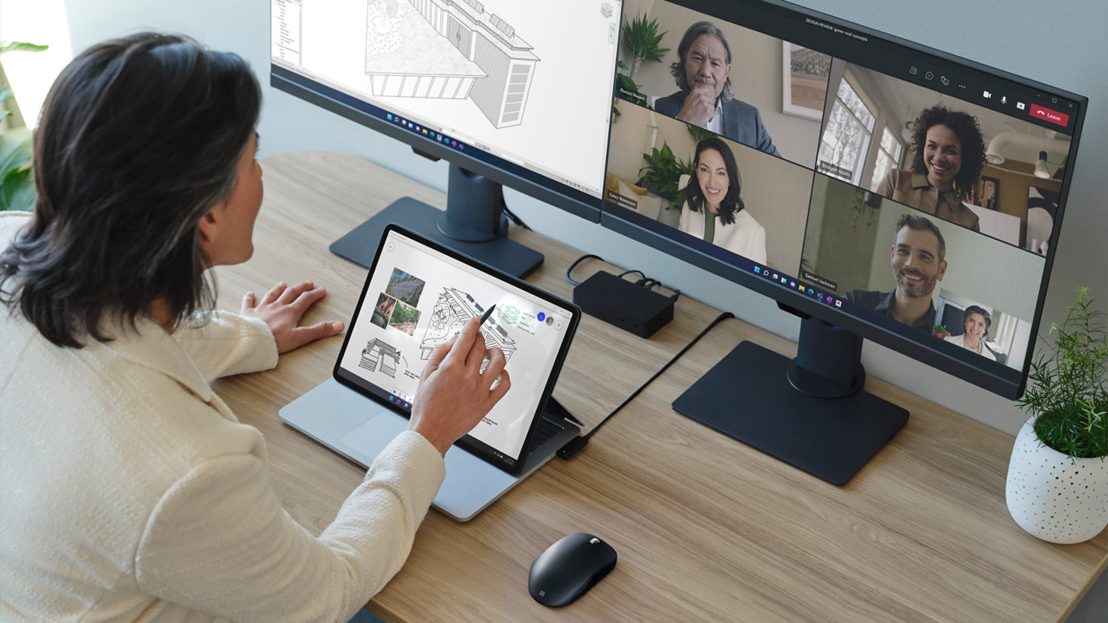 A person gesturing at the screen of Surface Laptop Studio for Business with a monitor in the background showing people in a Teams call.