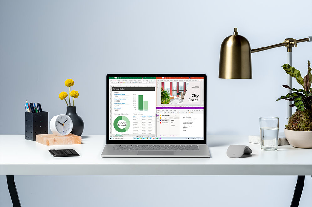 Front view of an open Surface laptop on a desk with lamp, plants, clock, and vase. 