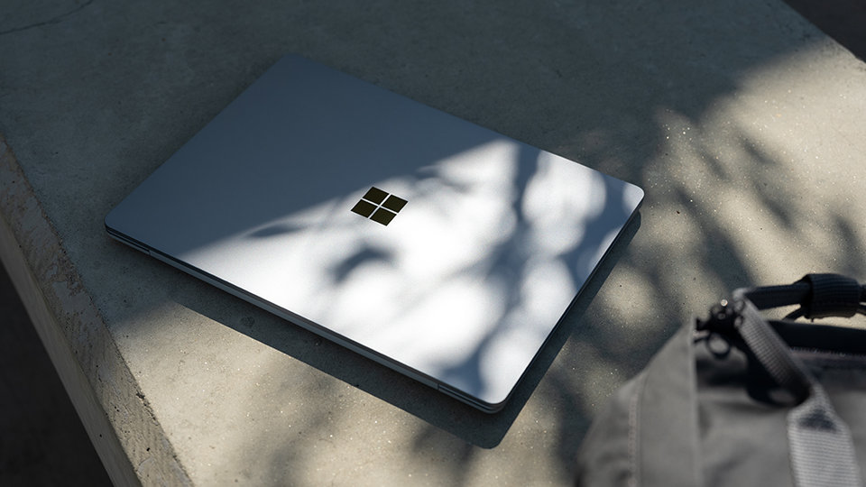 A Surface Laptop Go sits closed upon a concrete bench with a ruckpack nearby.