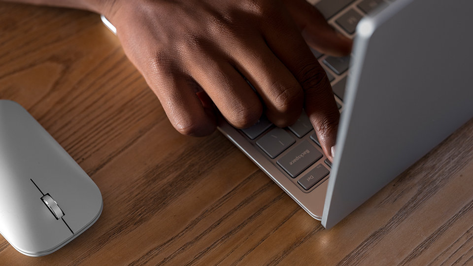 A person’s hand typing on the Surface Laptop Go keyboard with a Microsoft Modern Mobile Mouse nearby.