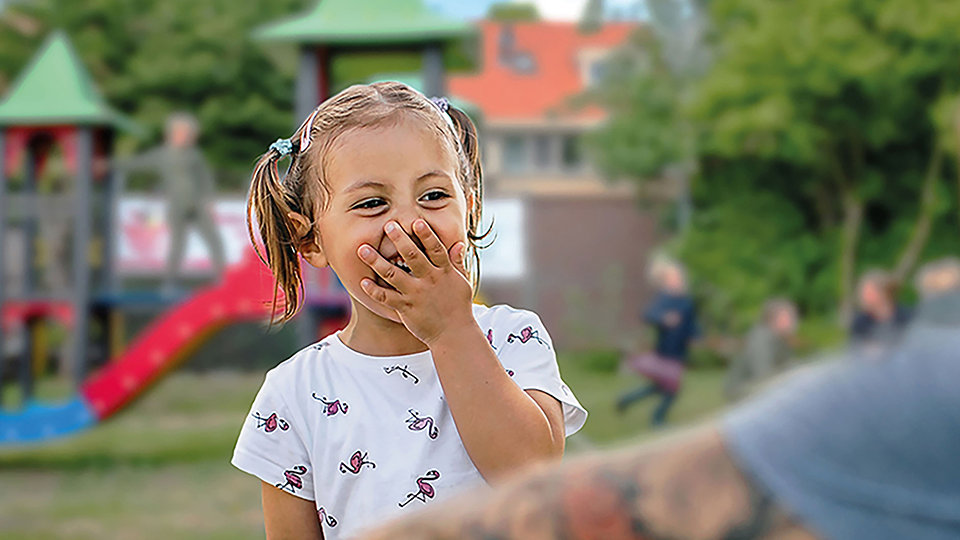 Child laughing with hand over mouth standing in front of a playground.