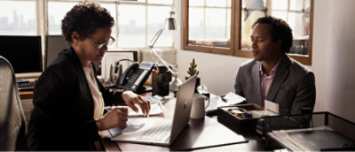 Two people at office desk with laptop and supplies.