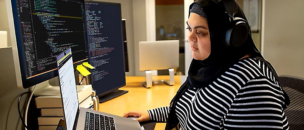 Eine Frau in einem schwarz-weiß gestreiften Hemd und Hijab, die an einem Computer arbeitet.