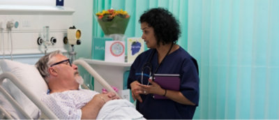 Medicinska sestra u uniformi drži tablet pored muškarca koji leži u bolničkom krevetu.