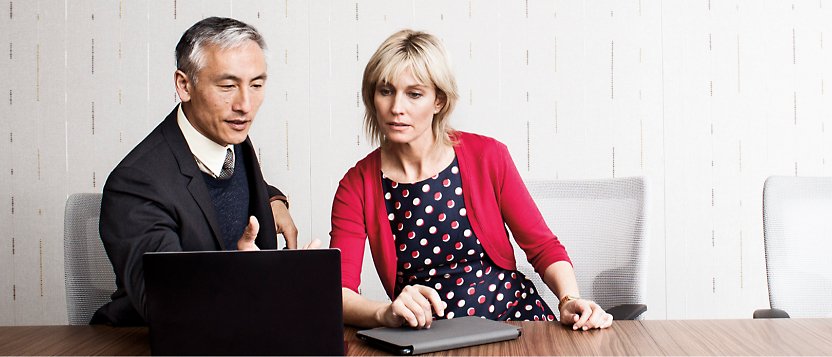 Un hombre y una mujer sentados en una mesa de conferencia miran un portátil.
